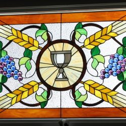 Stained glass window with a central chalice, grapes, wheat, green leaves, and geometric patterns.