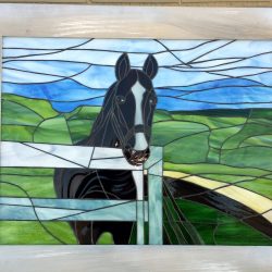 Stained glass artwork of a black horse standing by a white fence with green hills and blue sky in the background.