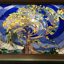Stained glass window featuring a tree with yellow leaves, set against a swirling blue and white background.