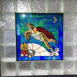Stained glass art of a sea turtle swimming with coral and bubbles, framed by textured clear glass blocks.