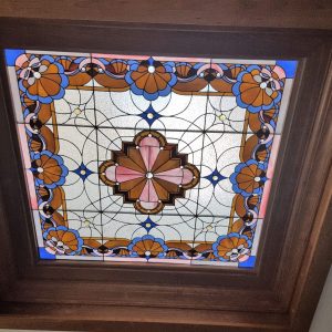 Stained glass ceiling panel with a geometric floral design in blue, pink, and brown tones, framed by dark wood.