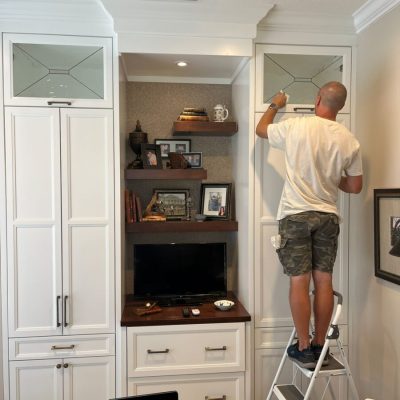 Man standing on a step stool cleans glass cabinet doors in a home office with white built-in shelves and a TV.