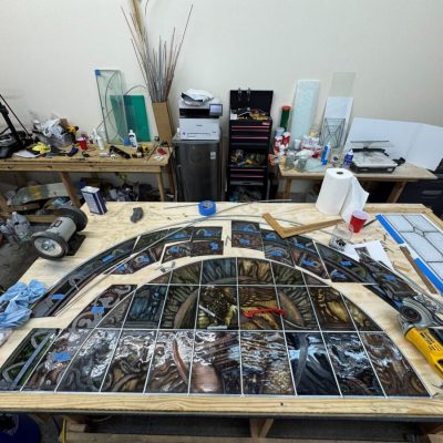 Large workbench with various stained glass pieces, tools, and materials arranged for an ongoing project in a workshop.