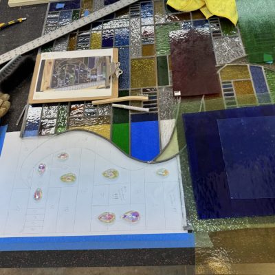 Colored glass pieces, gloves, a ruler, and a layout plan are arranged on a table for a stained glass project.