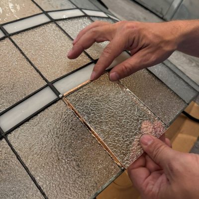Hands fitting a textured glass pane into a black-grid frame, assembling or repairing a stained glass window.