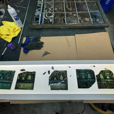 Broken stained glass pieces laid out on a white board, with tools and cleaning supplies visible on the workbench nearby.