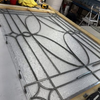 A decorative glass panel with metal framing rests on a workbench in a workshop with tools and supplies nearby.