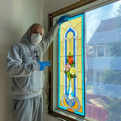 Person in gray hoodie, mask, and gloves installing or holding up a stained glass window panel indoors.