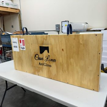 A wooden crate labeled “Casa Loma Art Glass” sits on a white table in a warehouse or workshop.