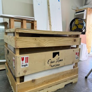Wooden crate labeled “Casa Loma Art Glass” with fragile stickers, placed in an indoor workspace with tools and equipment.