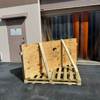 A large wooden shipping crate is positioned on a pallet outside a building with a closed roll-up door.