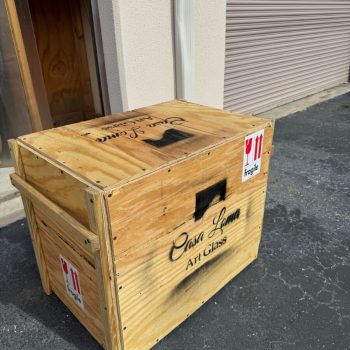Wooden crate labeled “Casa Loma Art Glass” with red “fragile” stickers sits on asphalt near a building.