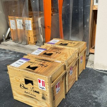 Three wooden crates labeled “Casa Loma Art Glass” sit on a paved surface near clear and orange plastic curtains.